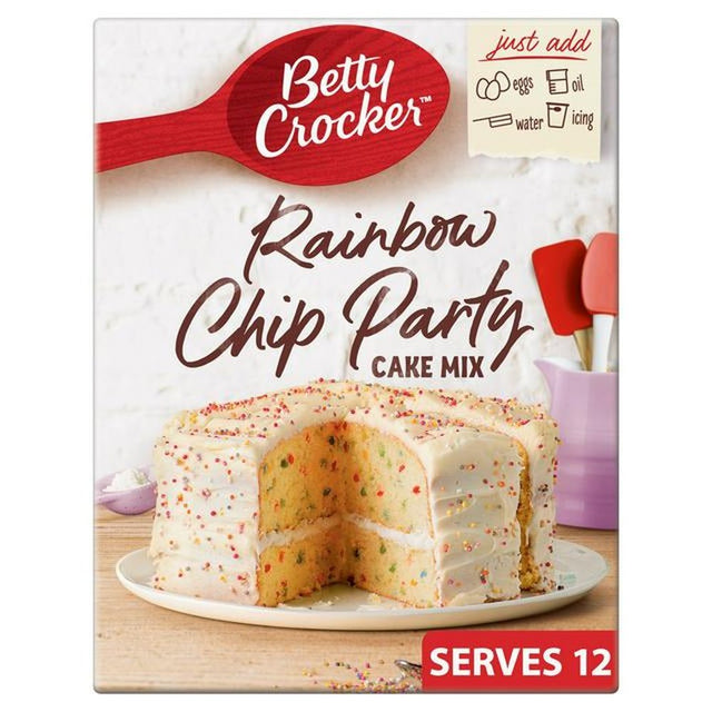 Un carton sur fond blanc avec un gâteau nature au glaçage à billes colorées sur une assiette blanche qui est sur une table en bois avec à l’arrière un mur blanc