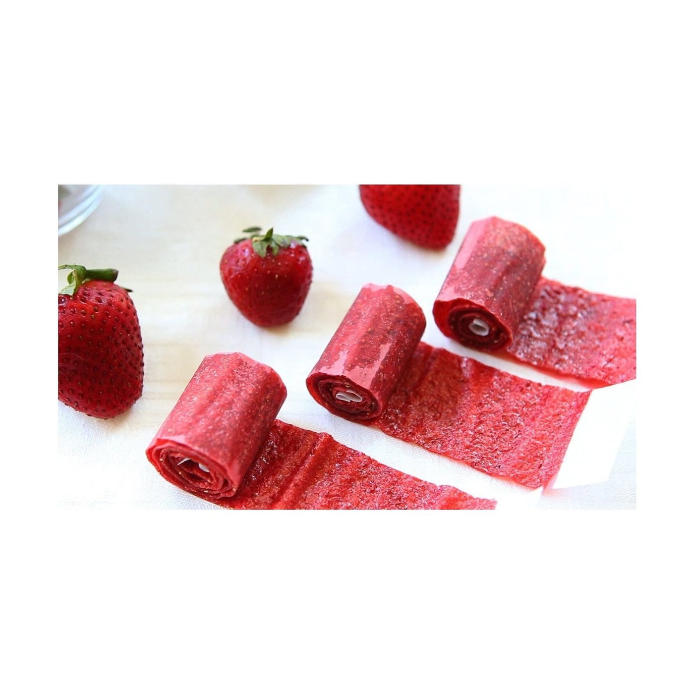 3 bonbons rouges enroulés devant 3 fraises sur une table avec une nappe blanche
