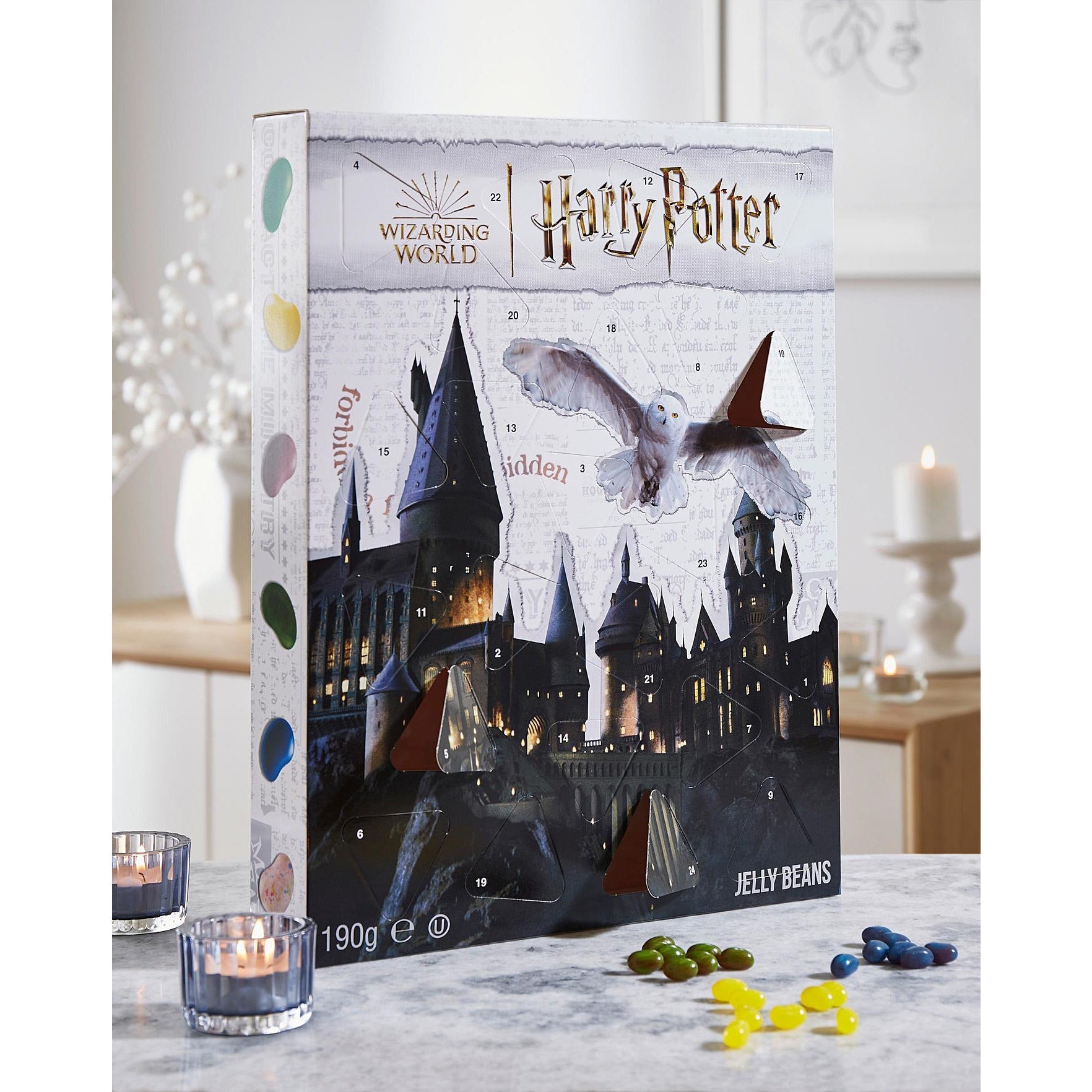 Le calendrier de l’Avent avec le château de Poudlard posé sur une table avec à gauche 2 bougies et à droite des Jelly Belly vert, jaune et bleu