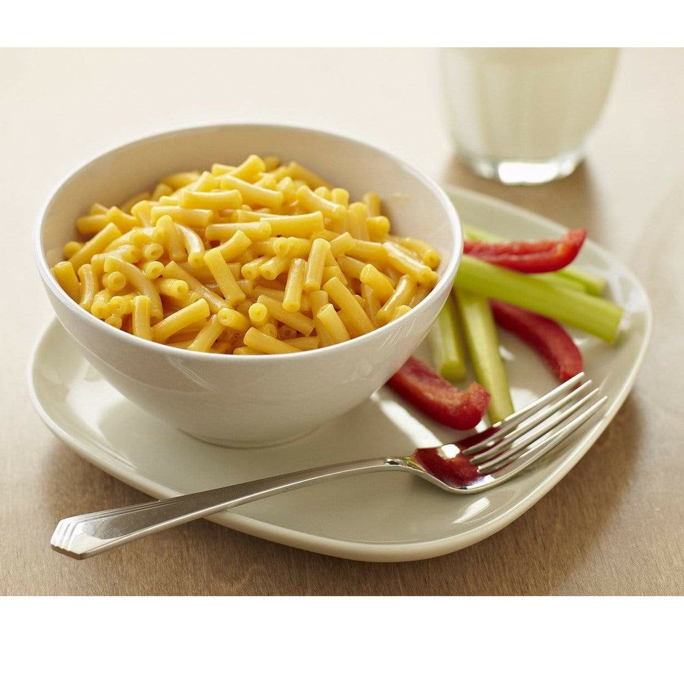 Un bol blanc de Mac&cheese sur une assiette blanche, sur le coté droit il y a du céleri et des poivrons coupés avec un fourchette