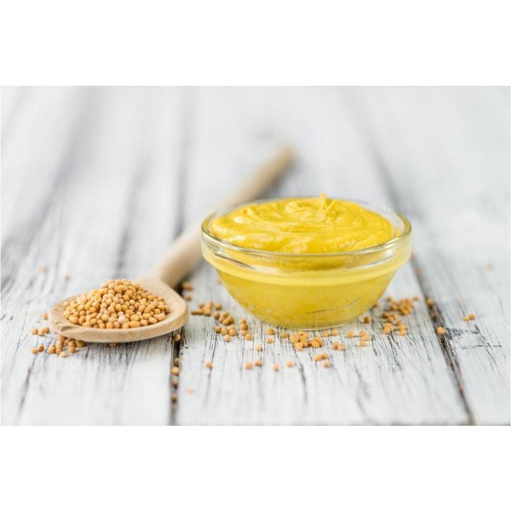 Un petit bol en verre rempli de de sauce jaune et à coté une cuillère en bois avec des graines de moutarde, le tout sur une table en bois gris