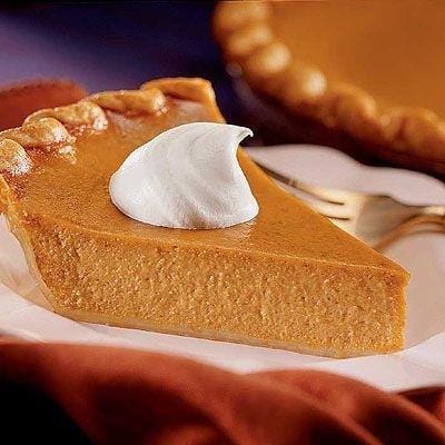 Une part de tarte orange avec de la chantilly blanche au-dessus, dans une assiette blanche avec un fourchette argenté. Le tout sur une nappe drapée orange