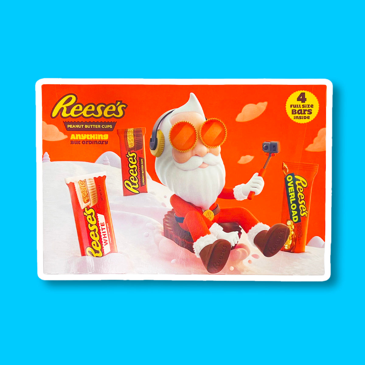 Un emballage orange sur fond bleu avec un père Noël stylé qui se filme entrain de descendre une montage enneigée assis sur un chocolat Reese’s géant et il y a 3 paquet de chocolat dans la neige