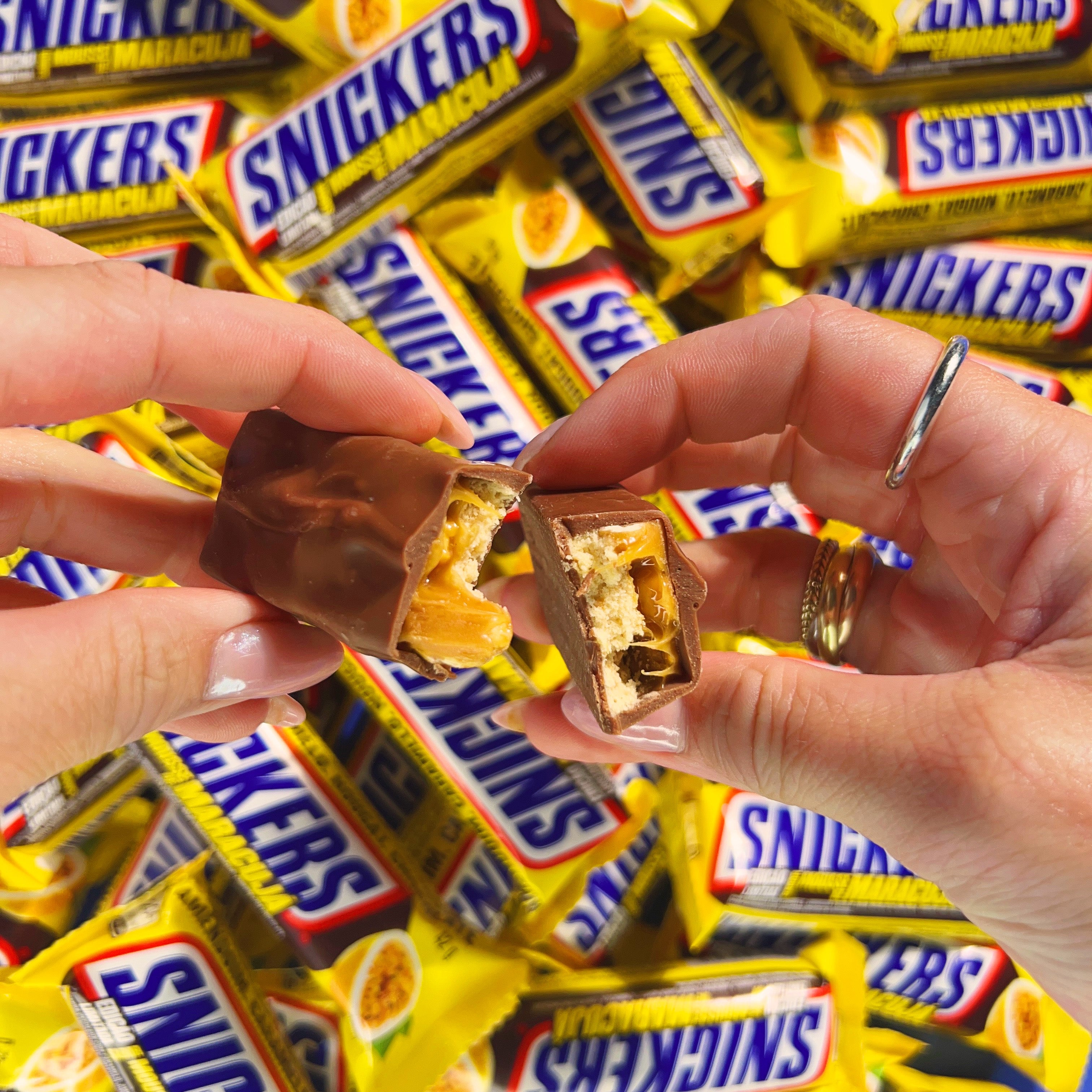 Une barre chocolatée coupée en 2 et tenue par 2 mains, à l'arrière il y a plein d'emballages jaunes de Snickers Maracuja