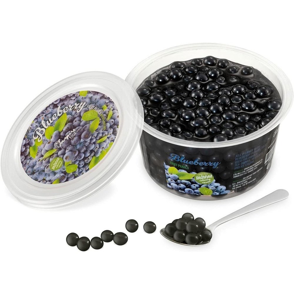 Une boite sur fond blanc remplie de perles noires et à gauche le couvercle avec plusieurs myrtilles. Devant des perles noires dans une cuillère