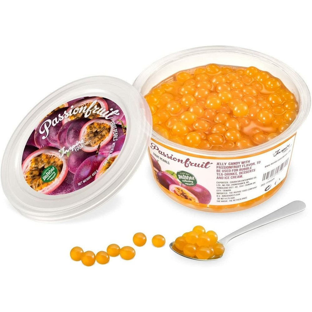 Une boite sur fond blanc remplie de perles oranges et à gauche le couvercle avec plusieurs fruits de la passion. Devant des perles oranges dans une cuillère