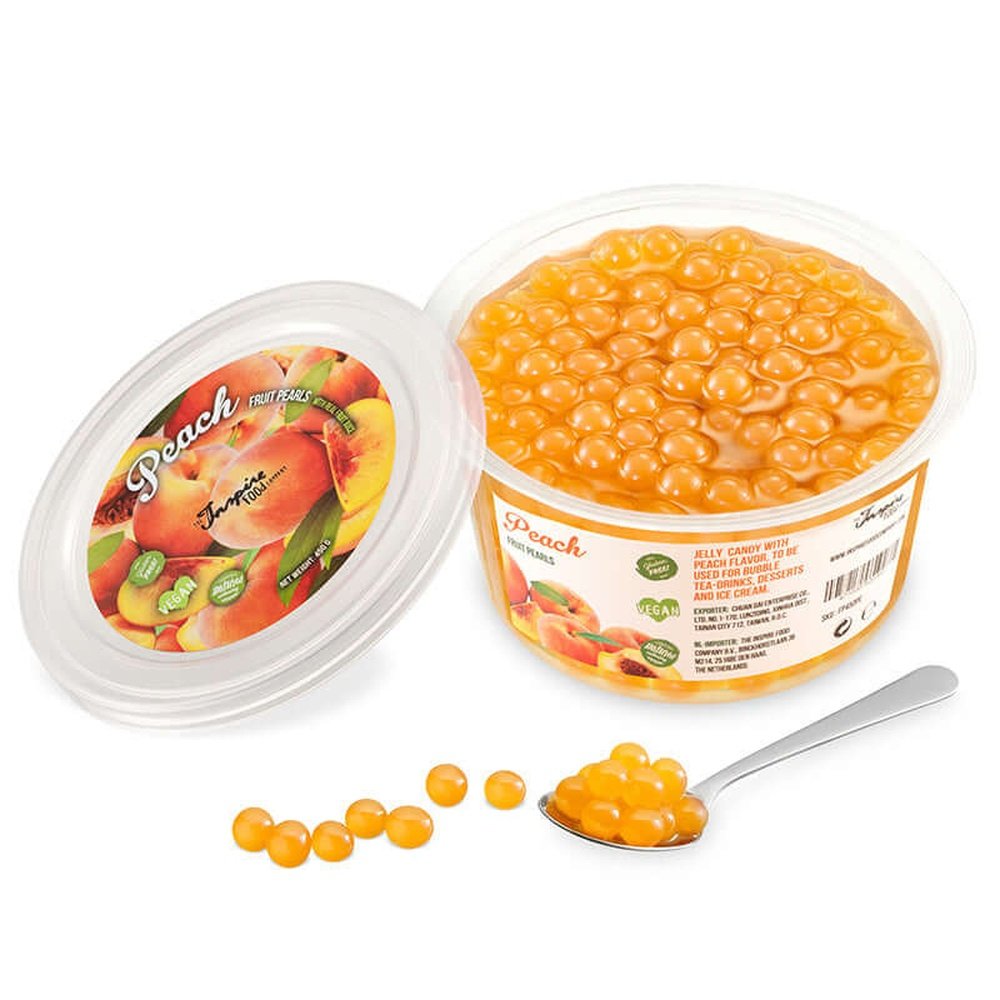 Une boite sur fond blanc remplie de perles oranges et à gauche le couvercle avec plusieurs pêches. Devant des perles oranges dans une cuillère