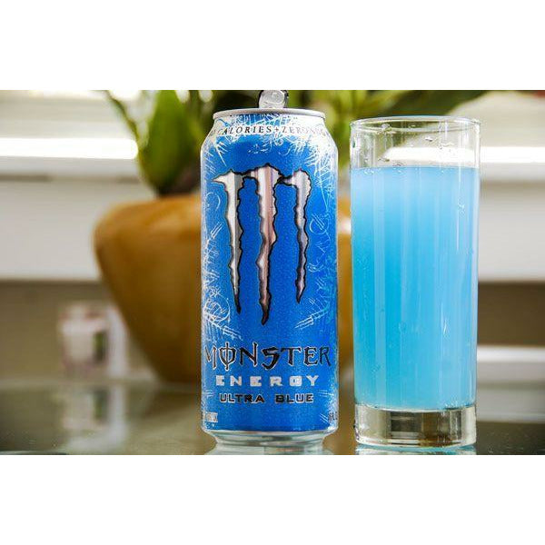 Une canette bleu à motifs argentés, au centre un M argenté de Monster et à côté un verre rempli d’une boisson bleu. Le tout sur une table en verre