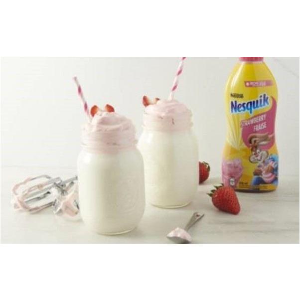 2 verres de lait avec de la mousse rose et à droite une bouteille jaune et rose, un capuchon rose. Le tout sur une table en bois clair avec des fraises