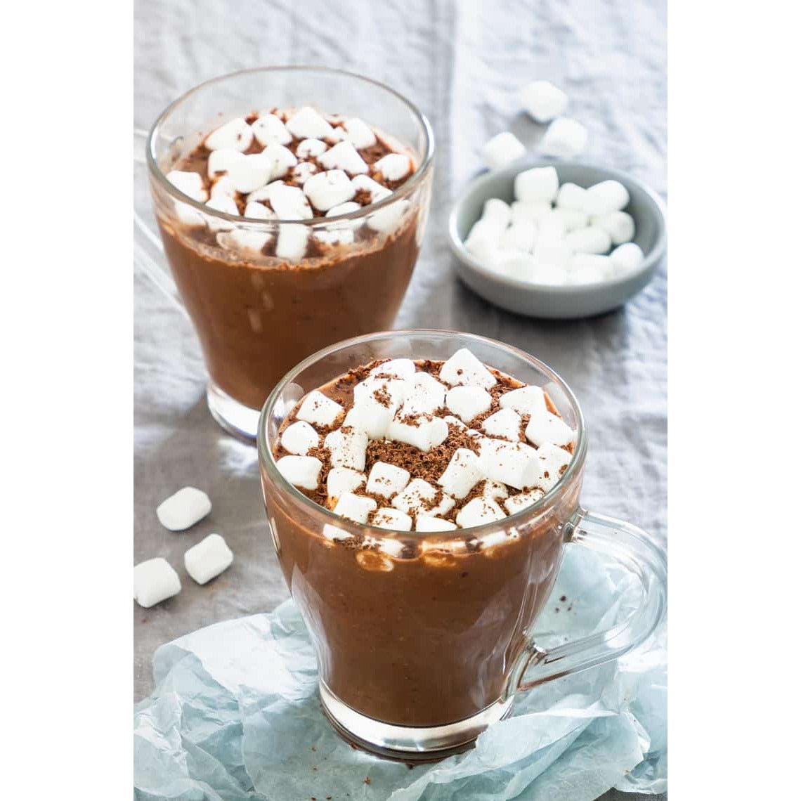 Deux tasses transparentes de chocolat chaud avec des mini marshmallows et derrière à droite un bol gris de marshmallows, le tout sur une nappe grise 