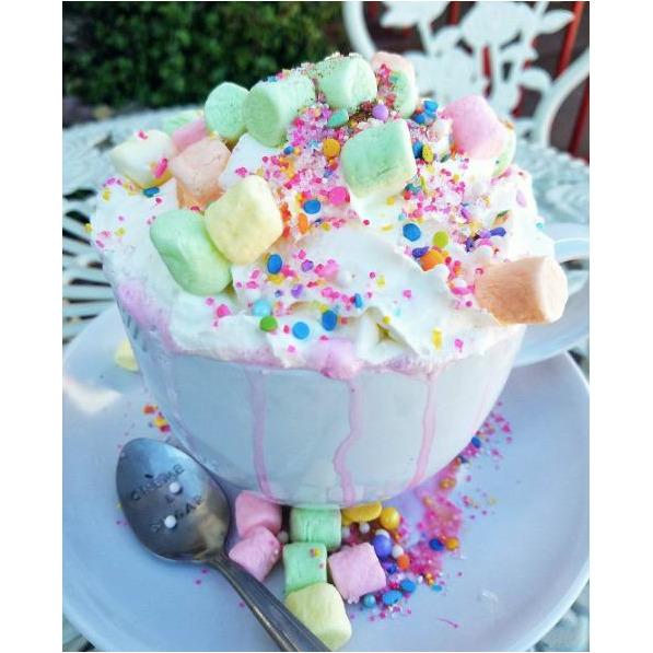 Une tasse blanche de chocolat chaud avec plein de crème chantilly qui dégouline et des marshmallows verts, jaunes et roses et plein de petites perles comestibles 