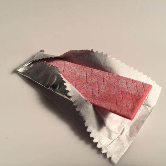 Un emballage individuel gris argenté qui d’ouvre et on y voit un chewing-gum rouge, sur une table blanche